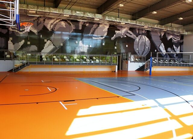 widok na salę sportowa wewnątrz boisko w kolorze pomaranczowo niebieskim z wyrysowanymi liniami, trybuny, kosze do koszykówki, ściana z grafiką przedstawiająca graczy grajacych w koszykówkę