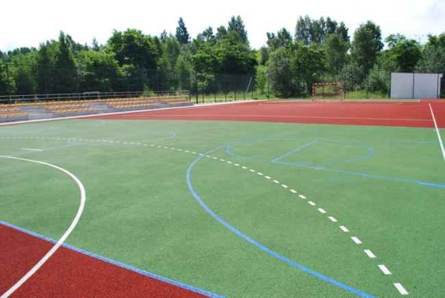 widok na płyte boiska z poliuretanu w kolorze bordowo zielonym z namalowanymi pasami wytyczajacymi boiska, w tle bramka oraz drzewa i krzewy po lewej str. trybuna sportowa