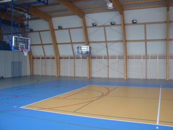 widok na hale, płyta boiska parkiet w kolorze jasnego drewna i nienbieskim, z narysowanymi liniami boisk do gier, siatka do siatkówki, kosz do koszykówki, po prawej stronie okna