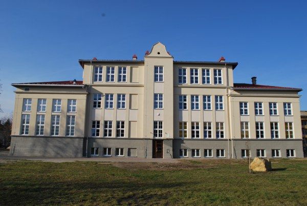 widok na tylna cześc budynku szkoły elewacja w kolorze ecru, parter w elewacja w kolorze ciemno szarym, 3 piętra w budynku płaski cimeno brązowy dach
