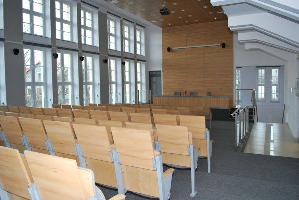 widok sali audiowizualnej, po lewej str ściana skaldająca się z dużych okien, miejsca siedzace w rzedach w kolorze jasnego drewna, dłówna lada w kolorze drewna za nia sciana drewniana z zawieszaonym ekranem rozwijanym lewa strona wykładzina szara