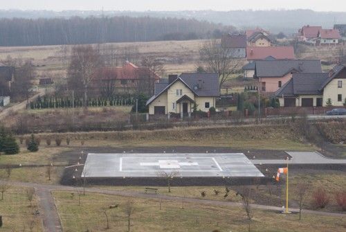 widok z lotu ptaka na lądowisko, płyta w kolorze szarym z białym znakiem krzża, wokó lasy, budynki mieszkalne, tereny zieleni - trwaniki