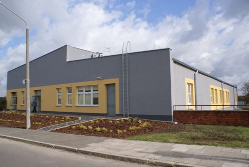 widok na budynek kliniki w kolorze szaro zółtym, budynek niski, przed nim zieleń i chodnik