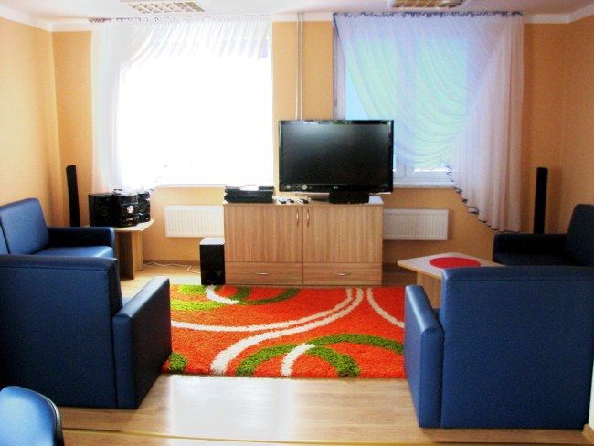 pokój z niebieskimi fotalami, pomarańczowym dywanem w pasy białe i zieone, szafka jasnobrązowa na niej telewizor, z tyłu okna z firankami, ścieny w kolorze jasny pomaranczowy