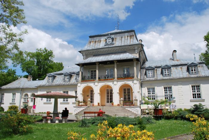 widok na zabytkowy pałac im. Starzeńskich w Płazie - obecnie siedziba dps budynek w kolorze jasny , okna , arkady przy wejsciu fłownym i kolumny przed budynkiem teren zielony trwawa  kwiaty krzewy i drzewa