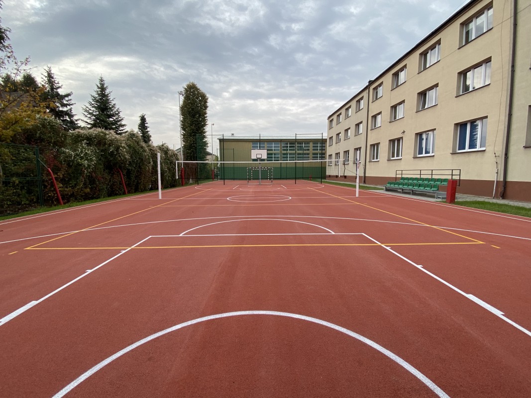 płyta boiska z narysowanymi liniami boisk, z siatką do siatkówki, koszem do koszykówki, po prawej stronie trybuna dla osób siedzących oraz budynek internatu w kolorze jasnym na wprost budynek hali sportowej w kolorze zółto - zielonym