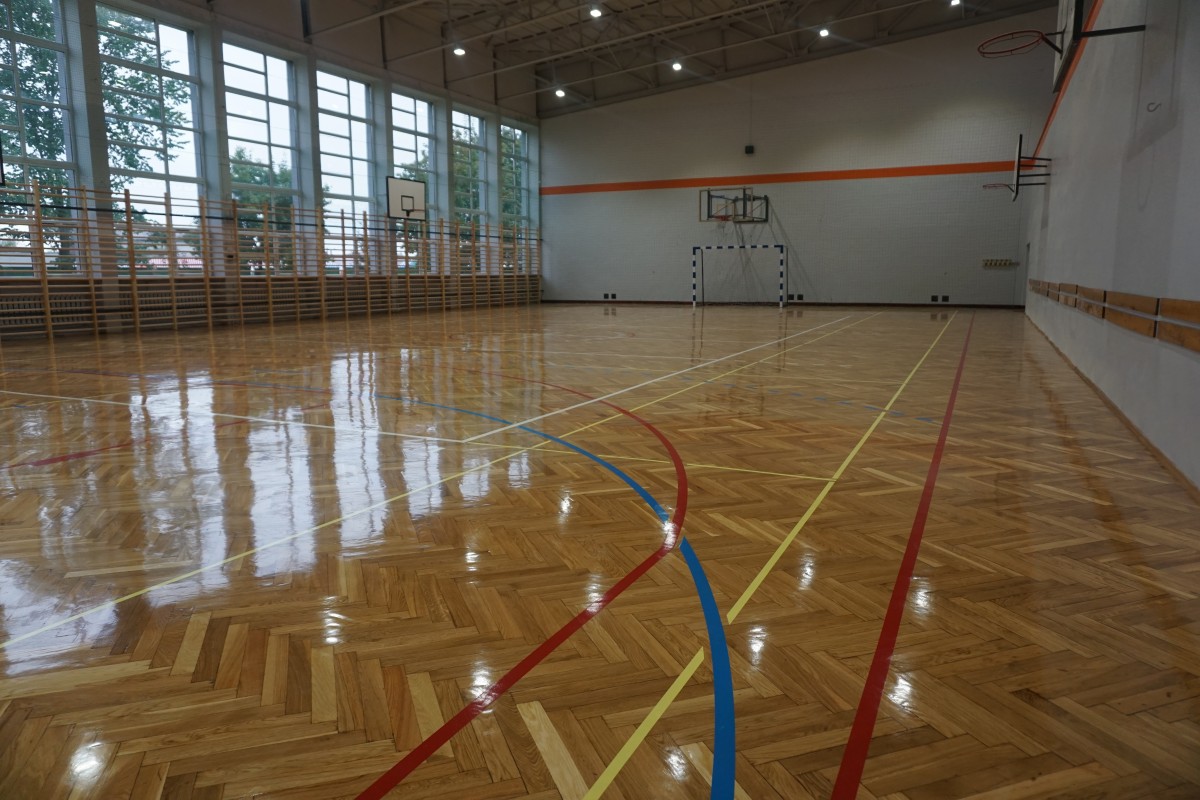 widok na wyremontowaną salę gimnastyczną nowy parkiet z narysowanymi polami boisk, kosze do koszykówki, bramka, biała ścana z pomaranczowym paskiem wzdłuz sciany, drabinki przy oknach