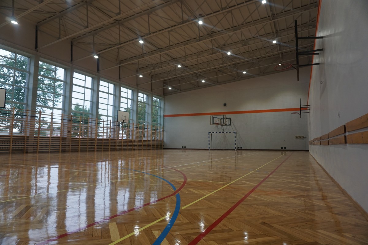 widok na wyremontowaną salę gimnastyczną nowy parkiet z narysowanymi polami boisk, kosze do koszykówki, bramka, biała ścane z pomaranczowum paskiem wzdłuz sciany, drabinki przy oknach