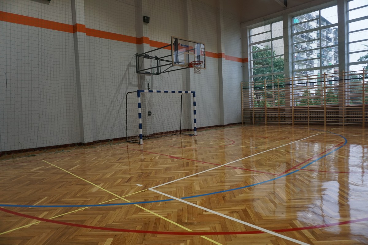 widok na wyremontowaną salę gimnastyczną nowy parkiet z narysowanymi polami boisk, kosze do koszykówki, bramka, biała ścane z pomaranczowum paskiem wzdłuz sciany
