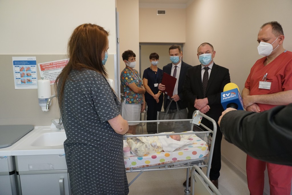 mama w szarej koszuli w jasne wzorki stoi przy łóżeczku niemowlaka, przed nią starosta chrzanowski, wicestarosta ubrani w garnitury, 2 panie z oddziału neonatologicznego oraz ordynator oddziału