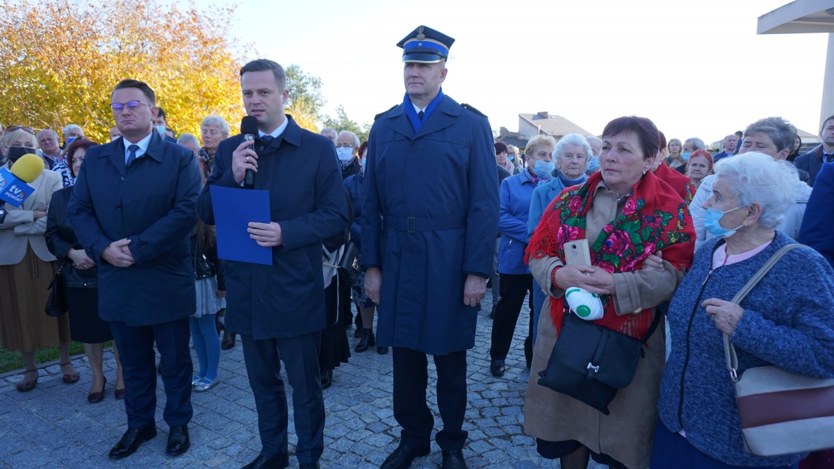 Mężczyzna w garniturze trzyma w ręce mikrofon. Przemawia. Obok zgromadzenie ludzi 