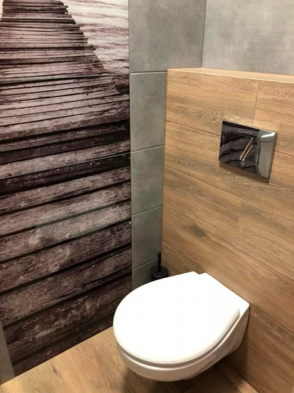 łazienka w wtz - toaleta- na wprost  scianka z widokiem na wode i kłądkę, po prawej str wc, wc zawieszone na ściance w kolorze drena jasnefo, widac kawałek szarej sciany z kafelków