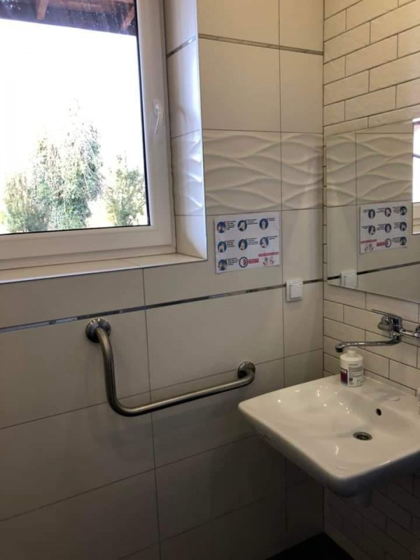 łazienka w wtz - po lewej str okno, pod oknem srebrny uchwyt dla niepełnosprawnych, po prawej umywalka  lustro, sciany białe z kafelek