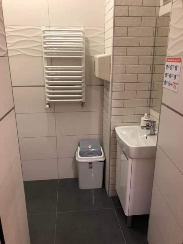 łazienka w wtz- na wprost kaloryfer pod nim j. szary kosz na śmieci po praej umywalka z szafką nad nią lustro, ściany białe z kafelek