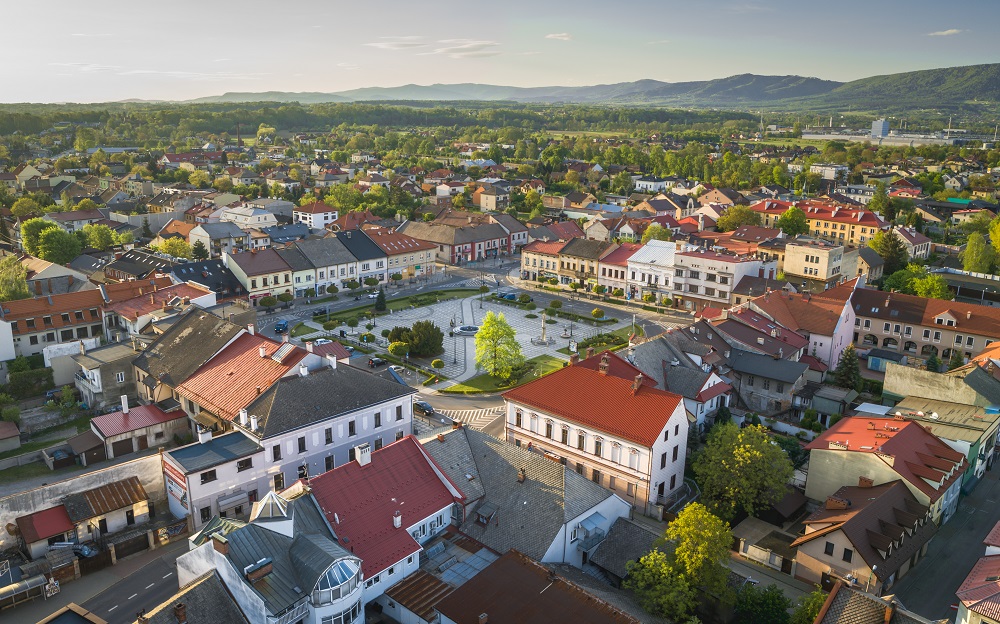 rynek w kętach na środku kwadratowy rynek wyłożony szarą kostką przeplatany zielenia i drzewami dookoła zarbudowania przyległych do siebie kamienic w tle widok na góry i tereny zielone