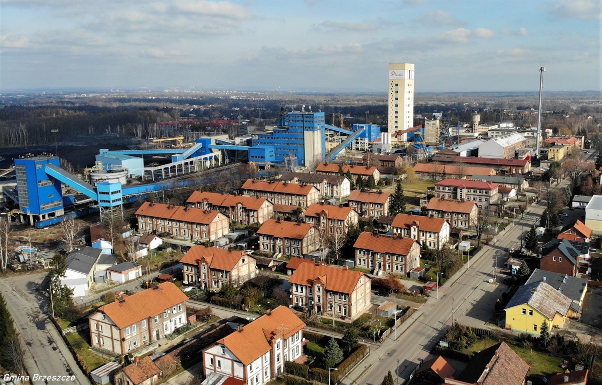 WIDOK NA OS. GÓRNICZE PO LEWEJ STRONIE ROZCIĄGAJA SIĘ NIEBIESKIE BUDYNKI KOPALNI, NA 1 PLANIE OSIEDLE BLOKÓW- 17 2-KONDYGNACYJNYCH W JASNO ŻÓŁTYCH Z RUDYM DACHEM, W TLE DRZEWA P PRAWEJ STRONIE PRZEBIEGA ULICA ASFALT SZARY, W ODDALI WYSOKI BUDYNEK ZÓŁTY