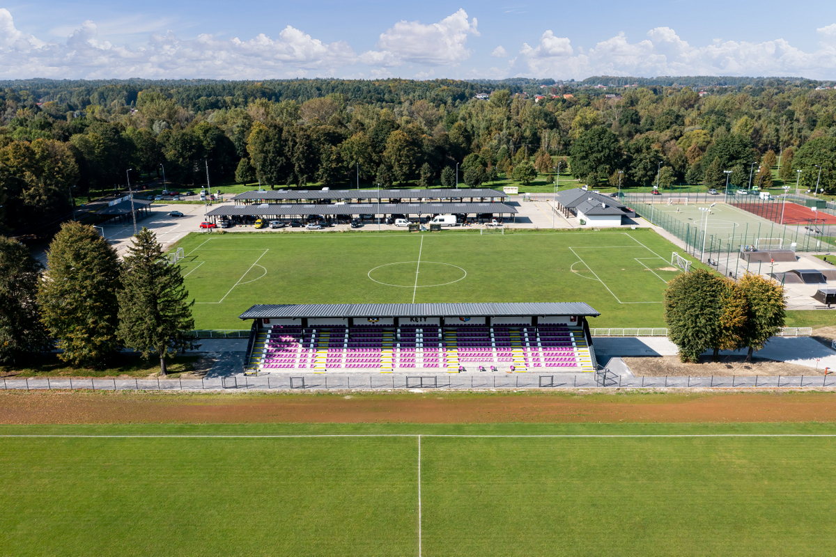 WIDOK NA STADION SPORTOW, MURAWA ZIELONA, TRYBUNY DA WIDZÓW, W ODDALI WIATY Z ZAPRAKOWANYMI AUTAMI,  PO PRAWEJ STR. BOISKA WIELOFUNKCYJNE  DO P. NOŻNEJ I O NAWIERZCHNI TARTANOWEJ NA WPROST W TLE ROŚLINNOSĆ, ZIELONE DZRWA I KRZEWY