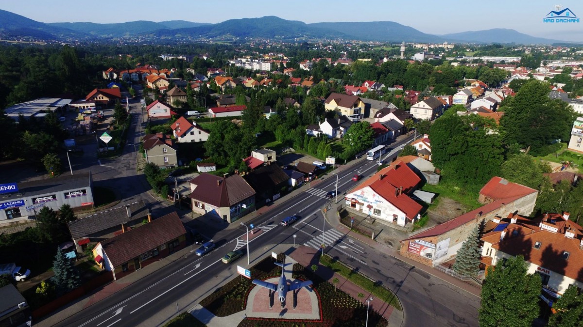 WIDOK NA MAISTO Z LOTU PTAKA, PO SRODKU ULICA ASFALTOWA NA ŚRODKU SKRZYZOWANIA SKWER Z KWIATAMI I RABATKAMI,  SAMOLOT,  PO BOKACH DROGI ZABUDOWNAIA LICZNE DOMY Z BRĄZOWYMI I RUDYMI DACHAMI, W TLE WIDOK NA GÓRY