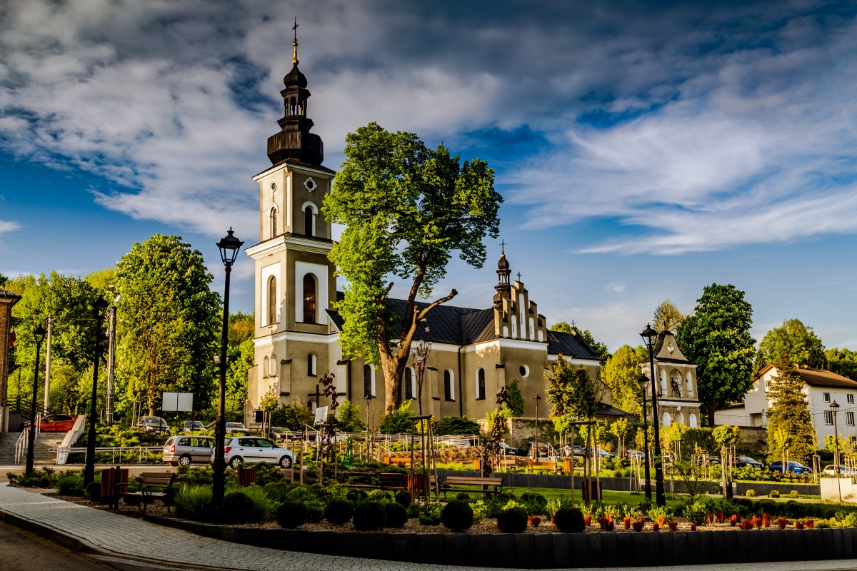 Szaro-biały budynek kościoła na tle zabudowąń, drzew i samochodów.