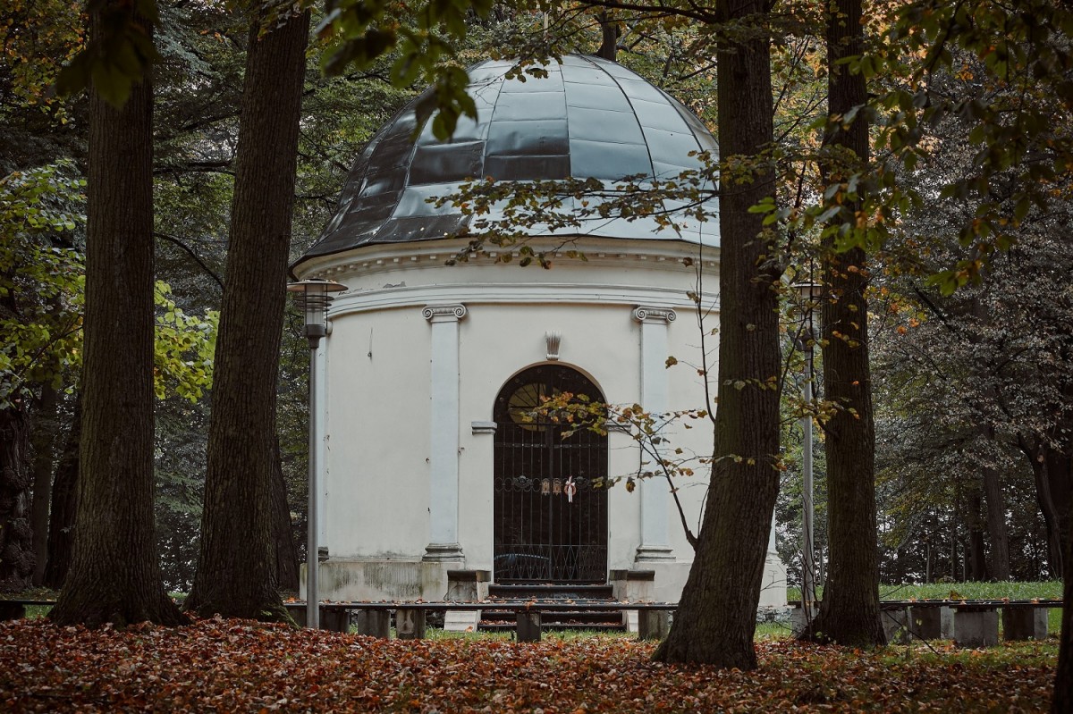 biała kaplica z kopułowatym dachem wokół drzewa i roślinność , przed kaplica ławki, dookoła rozsypane jesienne liście z drzew