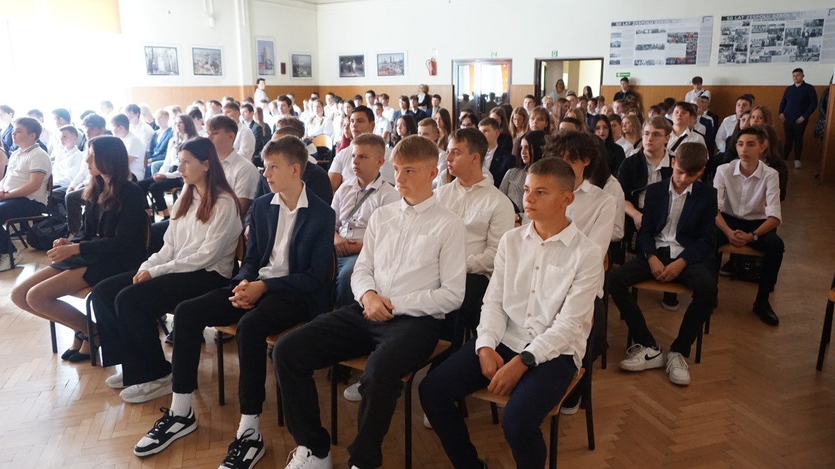 Młodzież ubrana w białe koszule i ciemne spodnie/spódnice siedzi obok siebie 