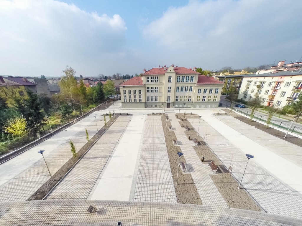 widok z lotu ptaka budynek pcku w chrzanowie w kolorze ecru z czerwonym dachem, duży parking z szarej kostki, pasy zielenie wzsłuż parkingu, lampy oświeylone po lewej stronie w tele budynki i drzewa po prawej stronie w tle budynki przy ul. focha