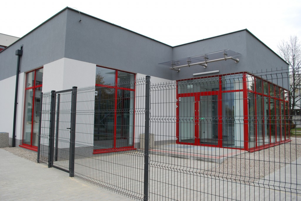 widok na budynek szatniowo administracyjny w kolorze jasno szarym , górna częśc elewacji w kolorze ciemno szraym ramy okien i dzrwi czerwone