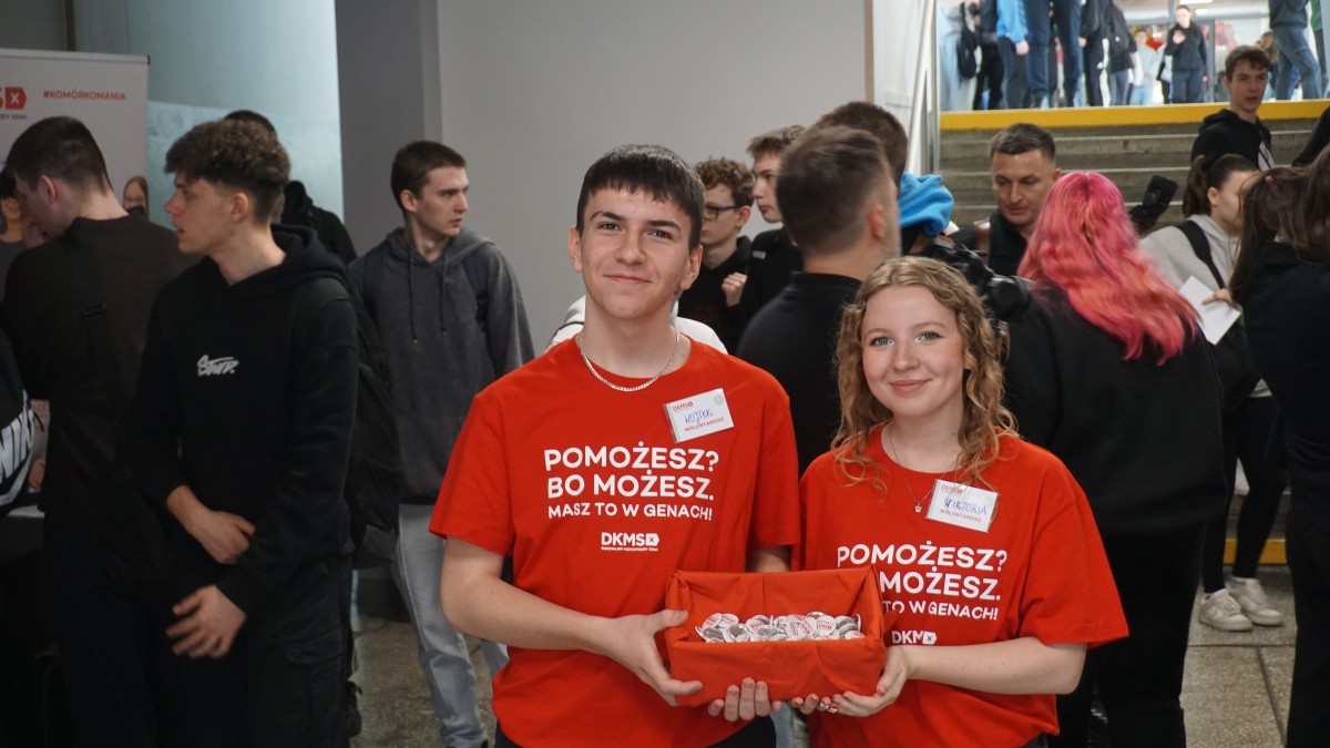 Zdjęcie dwóch wolontariuszy w czerwonych koszulkach tryzmających czerwone pudełko. W tle uczniowie szkoły.