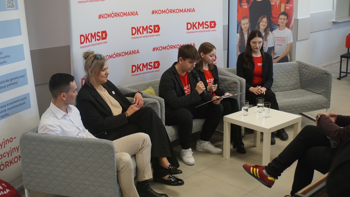 Zdjęcie 5 osób siedzących na kanapach przy stoliku. Jedna z nich trzyma mikrofon i przedstawia zasady panelu dyskusyjnego.
