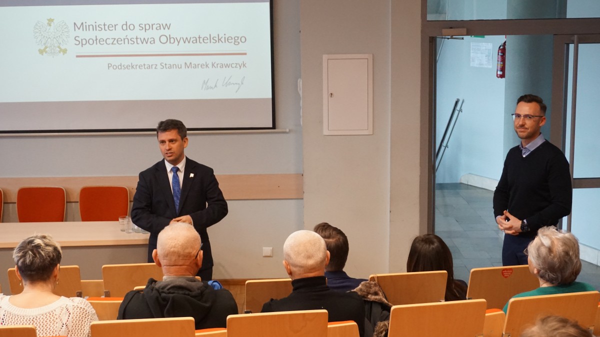 Zdjęcie kilku osób na auli. Dwójka z osób stoi frontem do osób siedzących na auli i do nich przemawia.