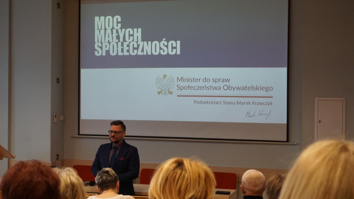 Zdjęcie kilku osób na auli. Jedna z osób stoi frontem do osób siedzących na auli i do nich przemawia, w tle rzutnik z prezentacją.