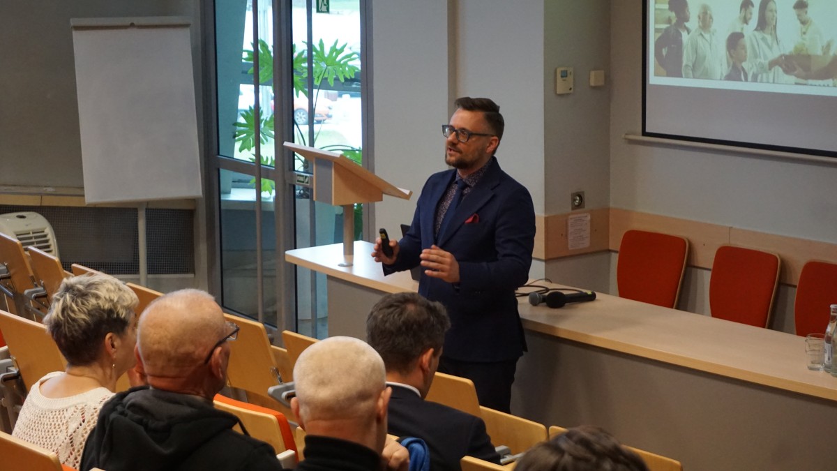 Zdjęcie kilku osób na auli. Jedna z osób stoi frontem do osób siedzących na auli i do nich przemawia.