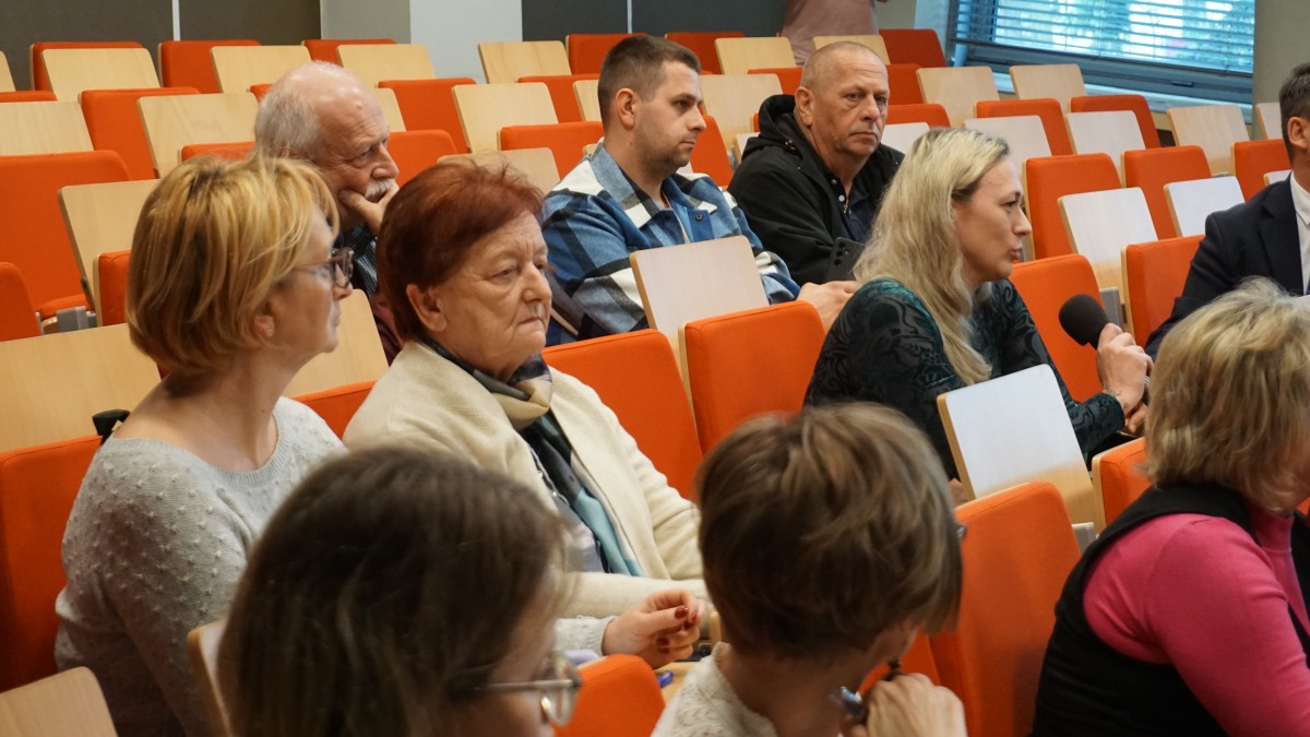 Zdjęcie kilku osób na auli. Wszystkie siedzą. Jedna z nich trzyma mikrofon i mówi.