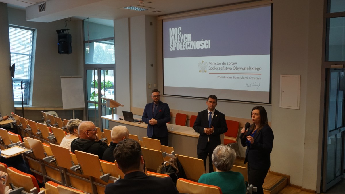 Zdjęcie kilku osób na auli. Trójka z osób stoi frontem do osób siedzących na auli i do nich przemawia.