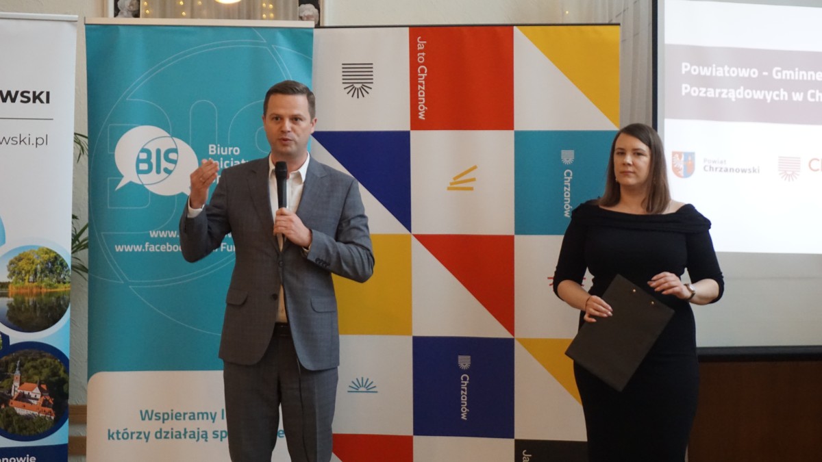 Zdjęcie dwóch osób, mężczyzny i kobiety. Mężczyzna stoi z lewej strony z mikrofonem i przemawia. Kobieta trzyma podkładkę pod karki. W tle roll'upy reklamowe organizatorów.