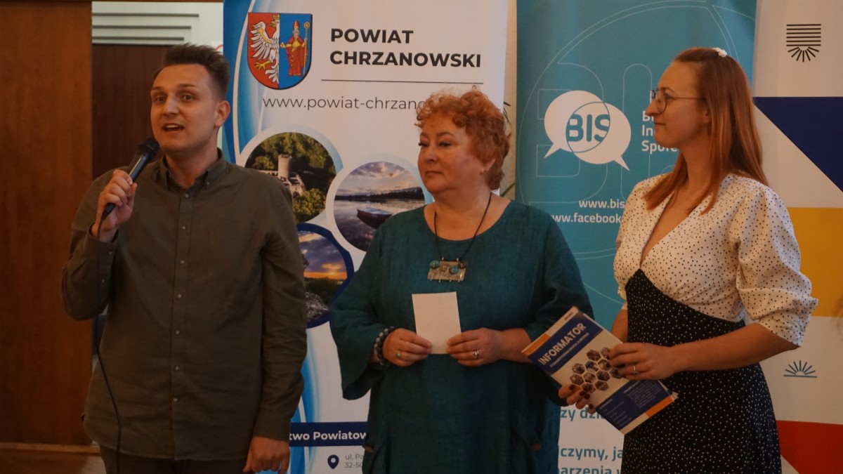Zdjęcie 3 osób stojących na środku sali. Jedna z osób trzyma mikrofon i przemawia do zebranych. W tyle roll'upy organziatorów.