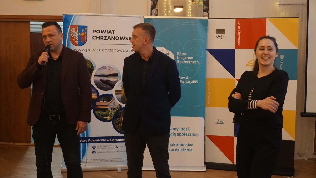 Zdjęcie 3 osób stojących na środku sali. Jedna z osób trzyma mikrofon i przemawia do zebranych. W tyle roll'upy organziatorów.