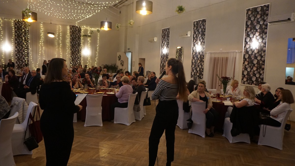Zdjęcie na którym dwie kobiety stoją na przeciwko osób na sali. Jedna z kobiet trzyma mikrofon i mówi do zebranych. Zebrani siedzą przy kilku okrągłych stołach. Przy każdym ze stołów siedzi kilkanaście osób.