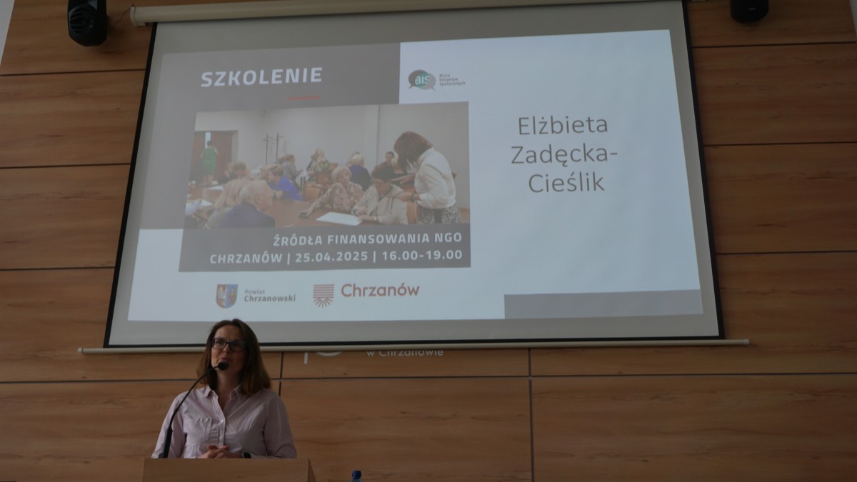 Kobieta stoi za pulpitem. Mówi do mikrofonu przed nią. Za nią jest rzutnik z prezentacją.