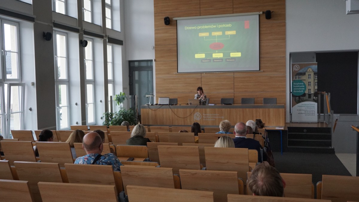 Zdjęcie kilkunastu osób siedzących na sali wykładowej. Większość siedzi i ogląda prezentacje wyświetlaną na rzutniku. Pod rzutnikiem vis-a-vis reszty osób siedzi kobieta z mikrofonem.