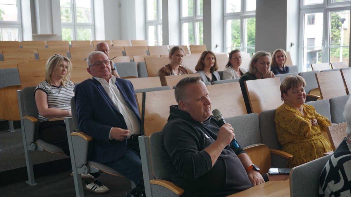 Zdjęcie dziesięciu osób w różnym wieku i różnej płci siedzących na sali wykładowej. Jedna z mężczyzn trzyma mikrofon i wypowiada się.