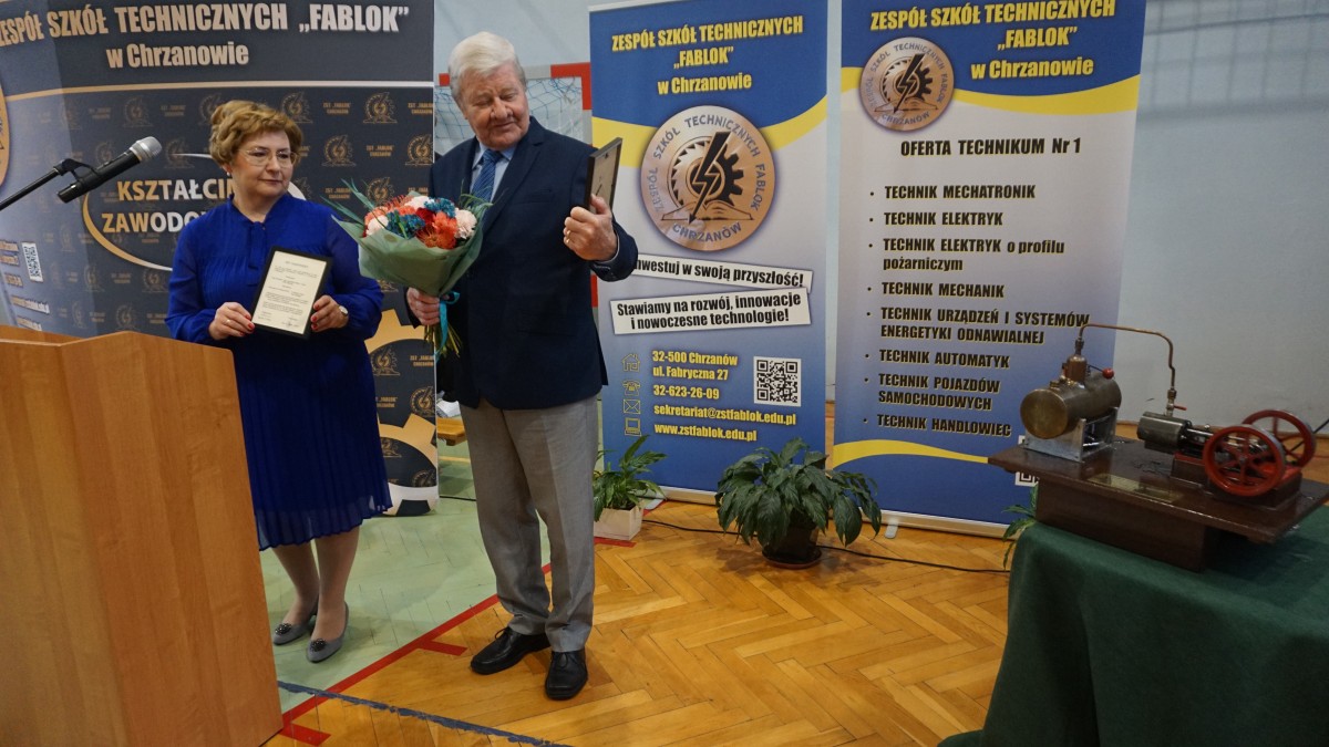 Kobieta i mężczyzna przy mównicy, nieopodal pierwowzór maszyny parowej