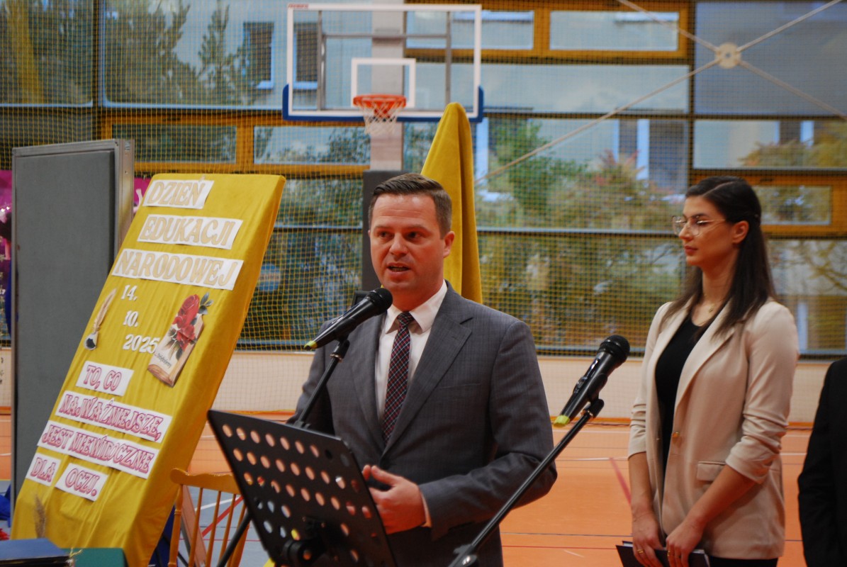 Zdjęcie mężczyzny w garniturze stającego za mikrofonem, na hali sportowej. W tle stoi młoda nauczycielka.