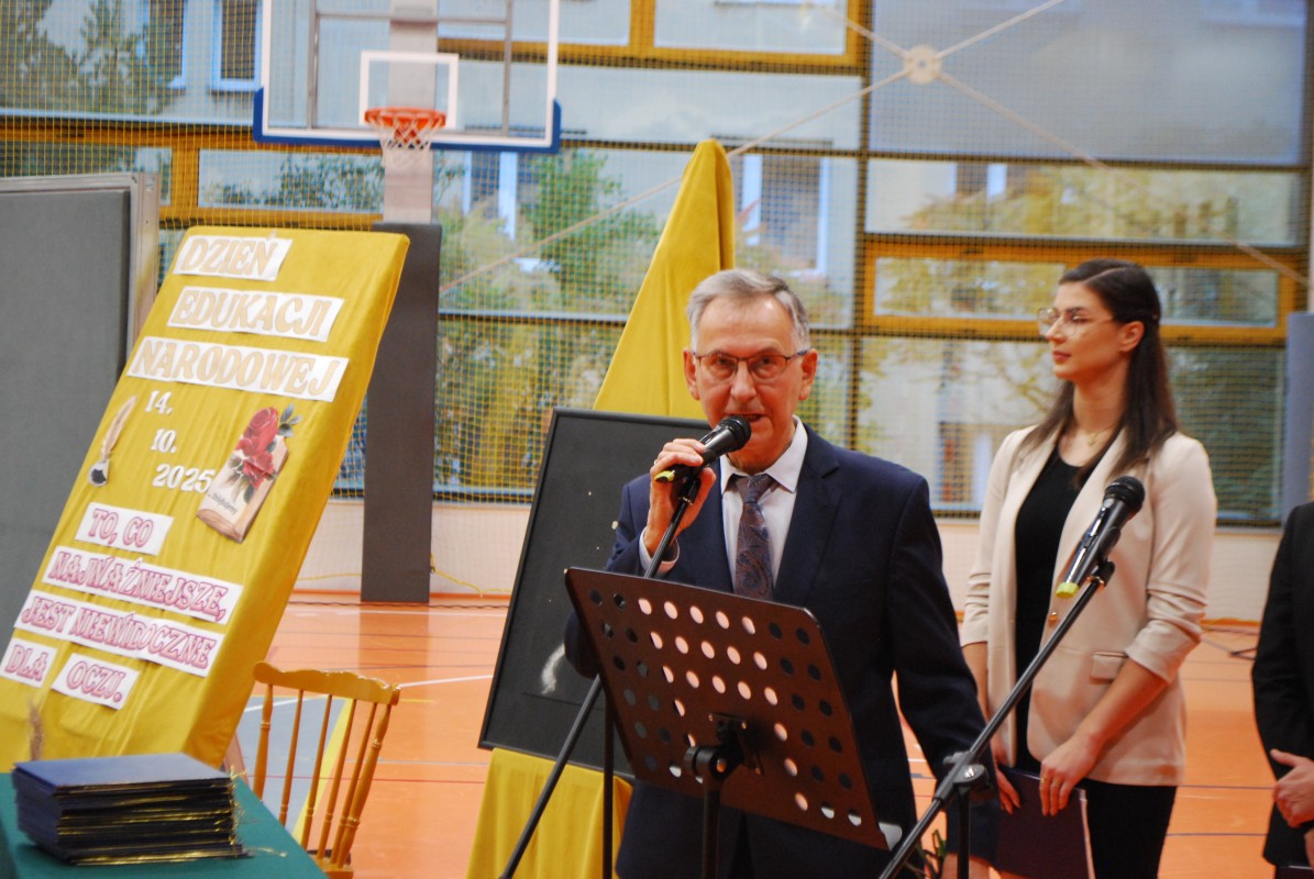 Zdjęcie mężczyzny w garniturze stającego za mikrofonem, na hali sportowej. W tle stoi młoda nauczycielka.