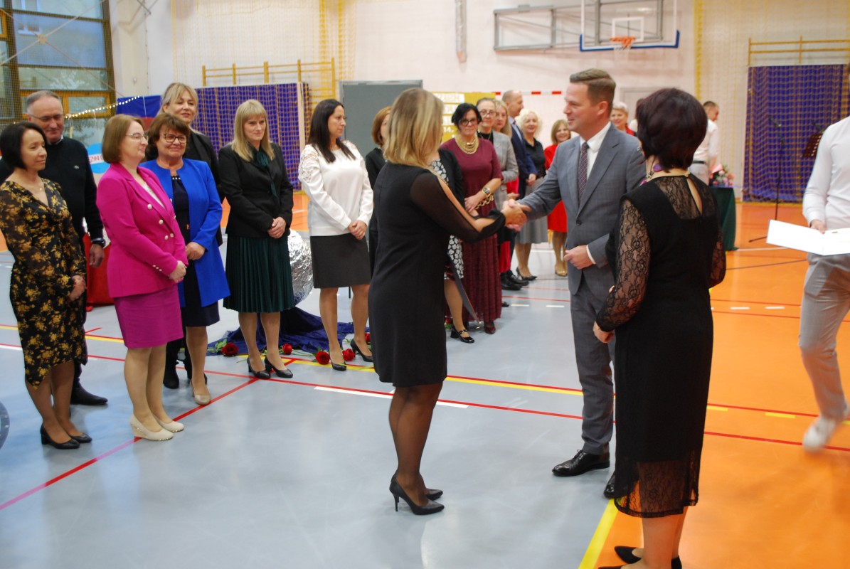 Kobieta i mężczyzna gratulują innej kobiecie. W tle grupa kilkudziesięciu innych nauczycieli.