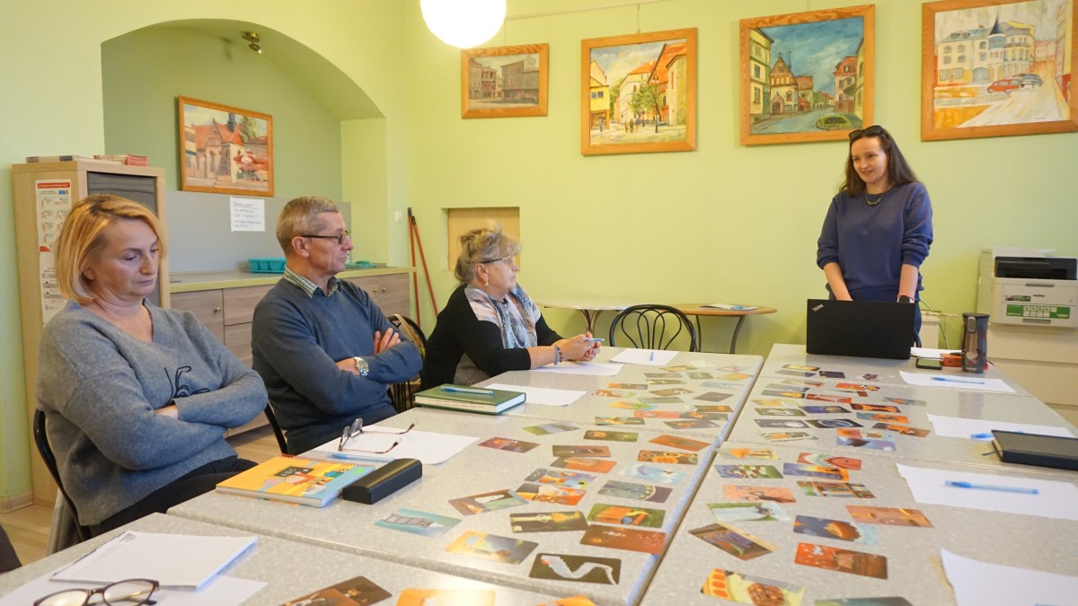 Zdjęcie czterech osób. Jedna osoba stoi przed nimi i mówi. Trzy osoby siedzą i słuchają.