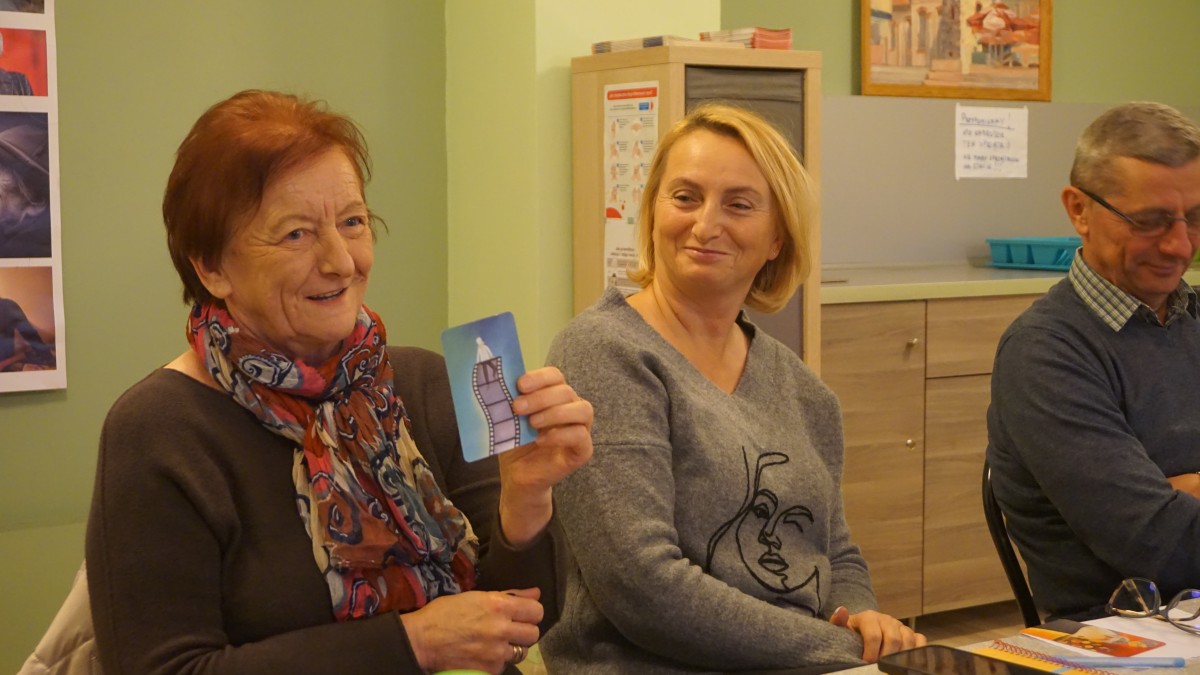 Zdjęcie mężczyzny i dwóch kobiet siedzących obok siebie. Jedna z kobiet trzyma kartę z obrazkiem.