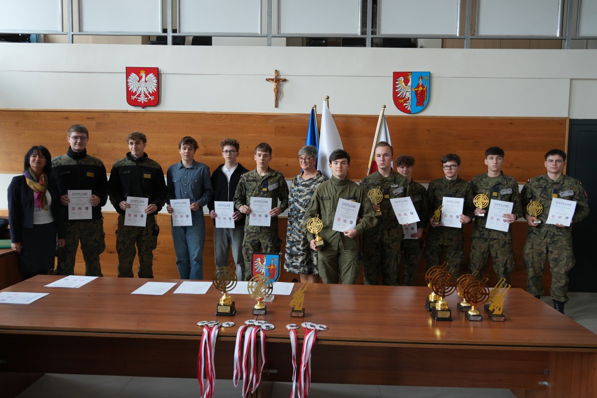 Grupa kilkunastu uczniów pozuje do zdjęcia z dwiema paniami. Uczniowe trzymają dyplomy oraz statuetki.