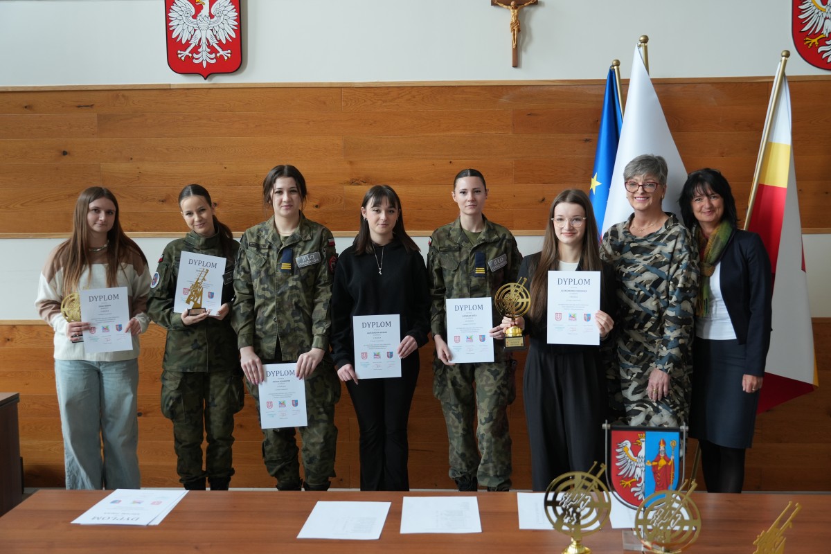 Grupa kilku uczniów pozuje do zdjęcia z dwiema paniami. Uczniowe trzymają dyplomy oraz statuetki.