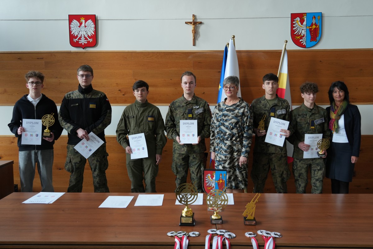 Grupa kilku uczniów pozuje do zdjęcia z dwiema paniami. Uczniowe trzymają dyplomy oraz statuetki.